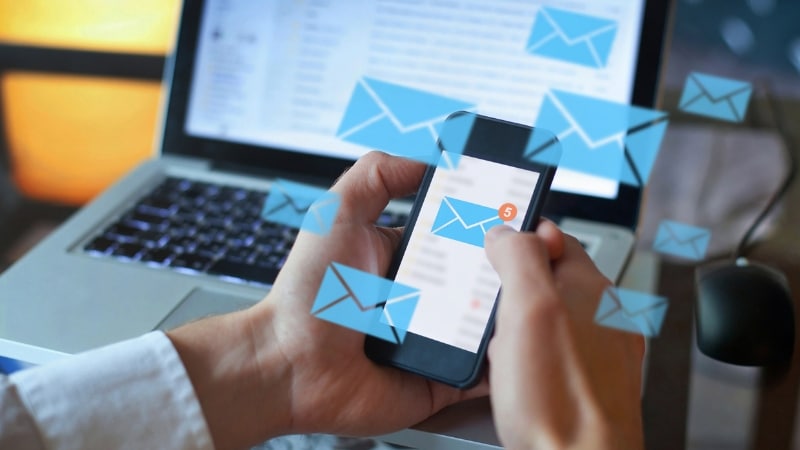A person checking emails on a smartphone with floating envelope icons, while a laptop is open in the background