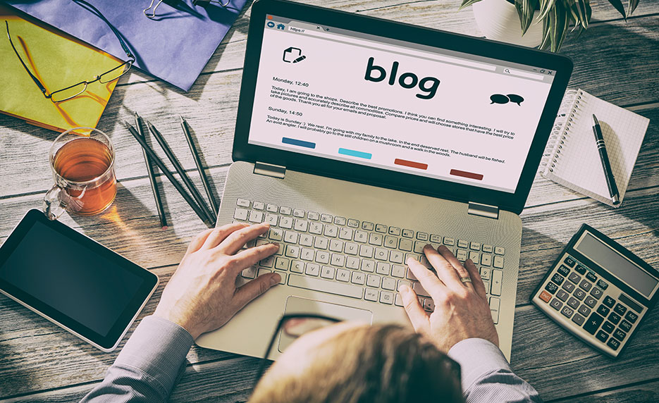 A person typing on a laptop with a blog open, surrounded by office supplies and a cup of tea