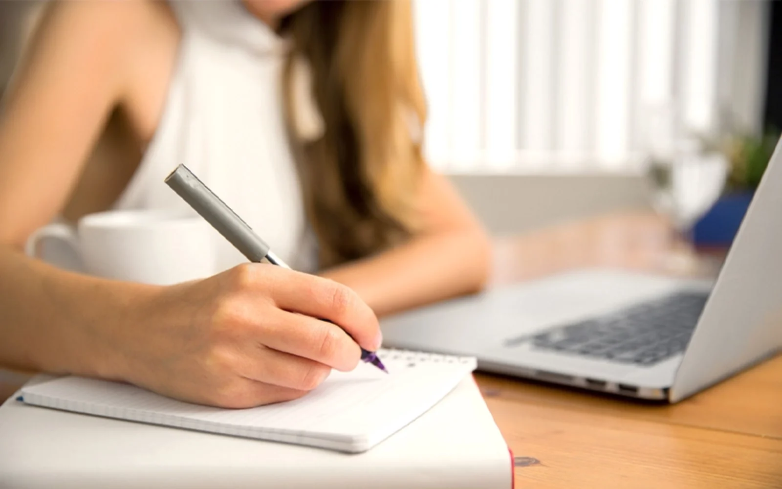 A person writing in a notebook with a pen while sitting at a desk with a laptop and a cup of coffee