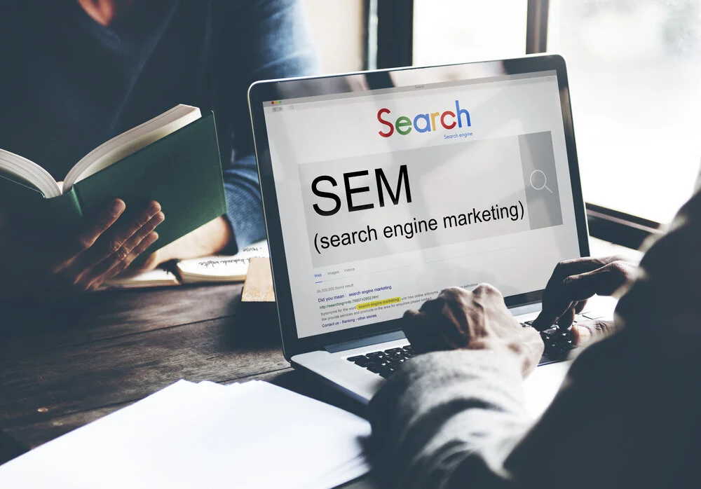 Person typing on a laptop searching for 'SEM (search engine marketing)' while holding a book.