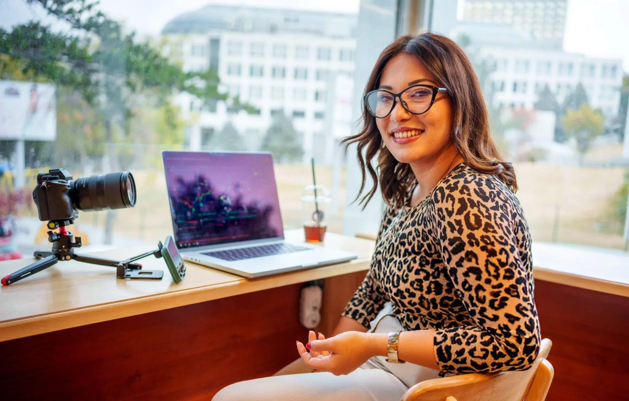 A woman smiling and sitting at a desk with a camera, laptop, and a drink in a well-lit space.