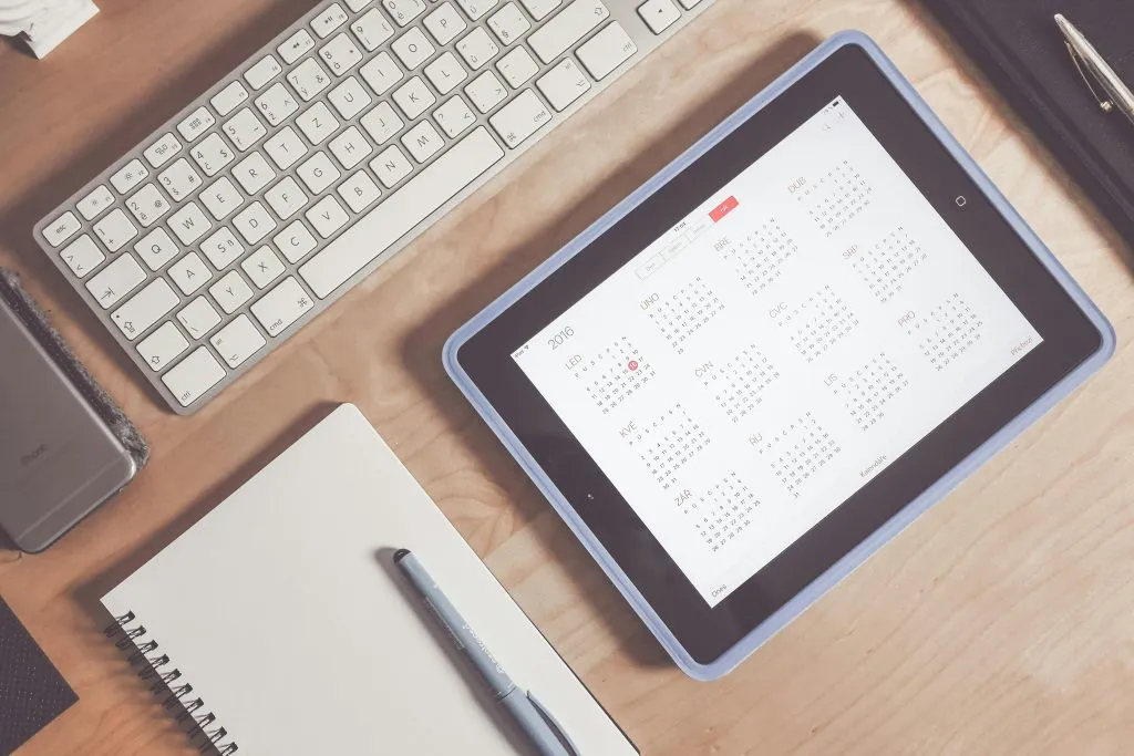 An overhead view of a desk with a keyboard, tablet displaying a calendar, notebook, and pen.