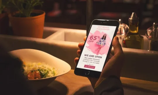 Hand holding a smartphone showing a summer sale advertisement while dining at a restaurant.