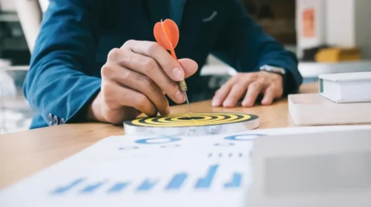 Person placing a dart in the bullseye of a target on a desk.