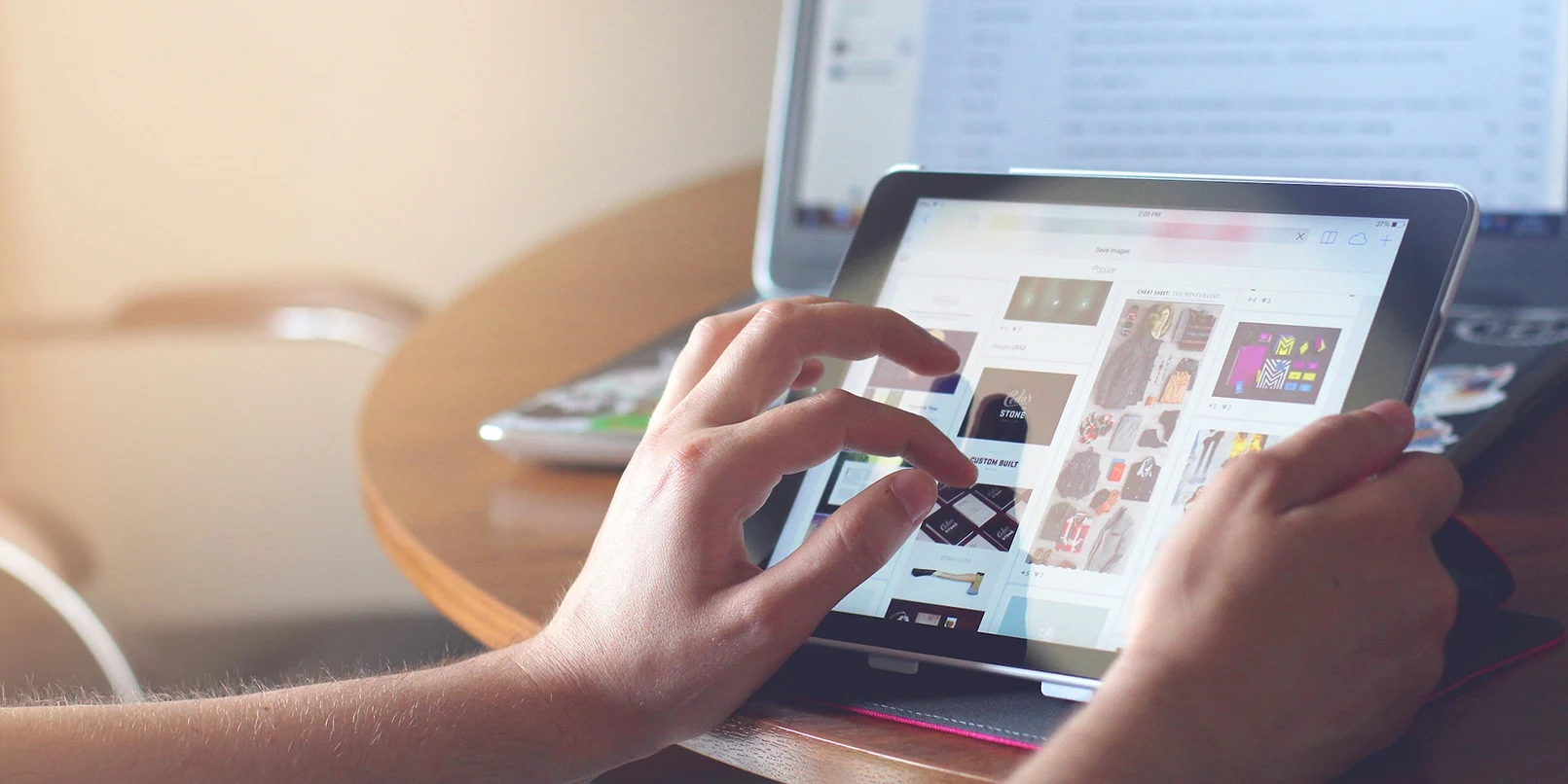 Someone browsing visual content on a tablet with a laptop in the background.