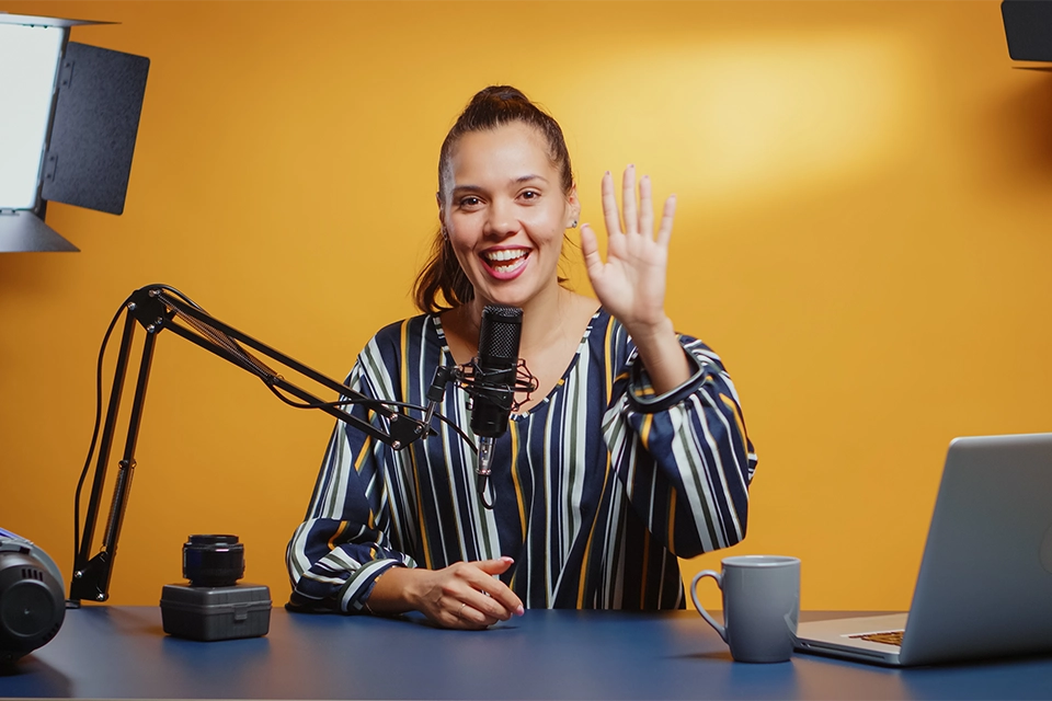 Woman recording a podcast while waving at a microphone in a studio setup.